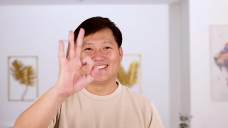 asian man smiles and raises his hand to make an okay gesture in a bright, modern indoor setting with soft, even lighting
