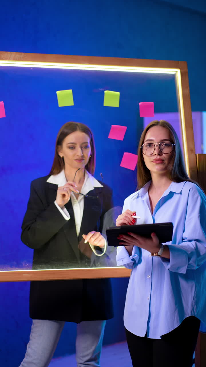 Smiling Caucasian ladies look at the tablet. Women working in the office stand at the glass wall with stickers. Vertical video.