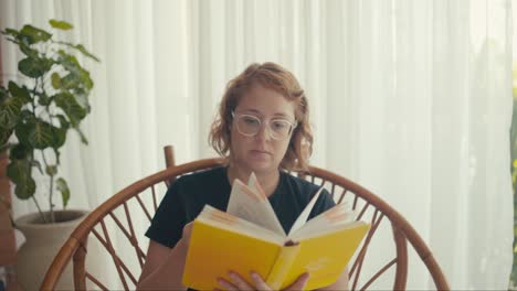 mujer de cabello rojo de mediana edad leyendo un libro sentada en una silla en su casa iluminada