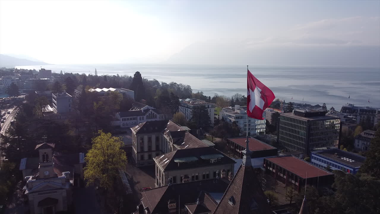 toma aérea lenta de la bandera suiza en la parte superior de un edificio renacentista en lausana, suiza en un día soleado con el lago de ginebra en el fondo