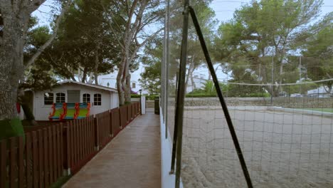 un dron se desliza paralelo a un patio de recreo infantil adornado con casas en miniatura y un parque de árboles, y a lo largo de un campo tanto de voleibol como de fútbol en el vibrante lugar de mallorca, españa