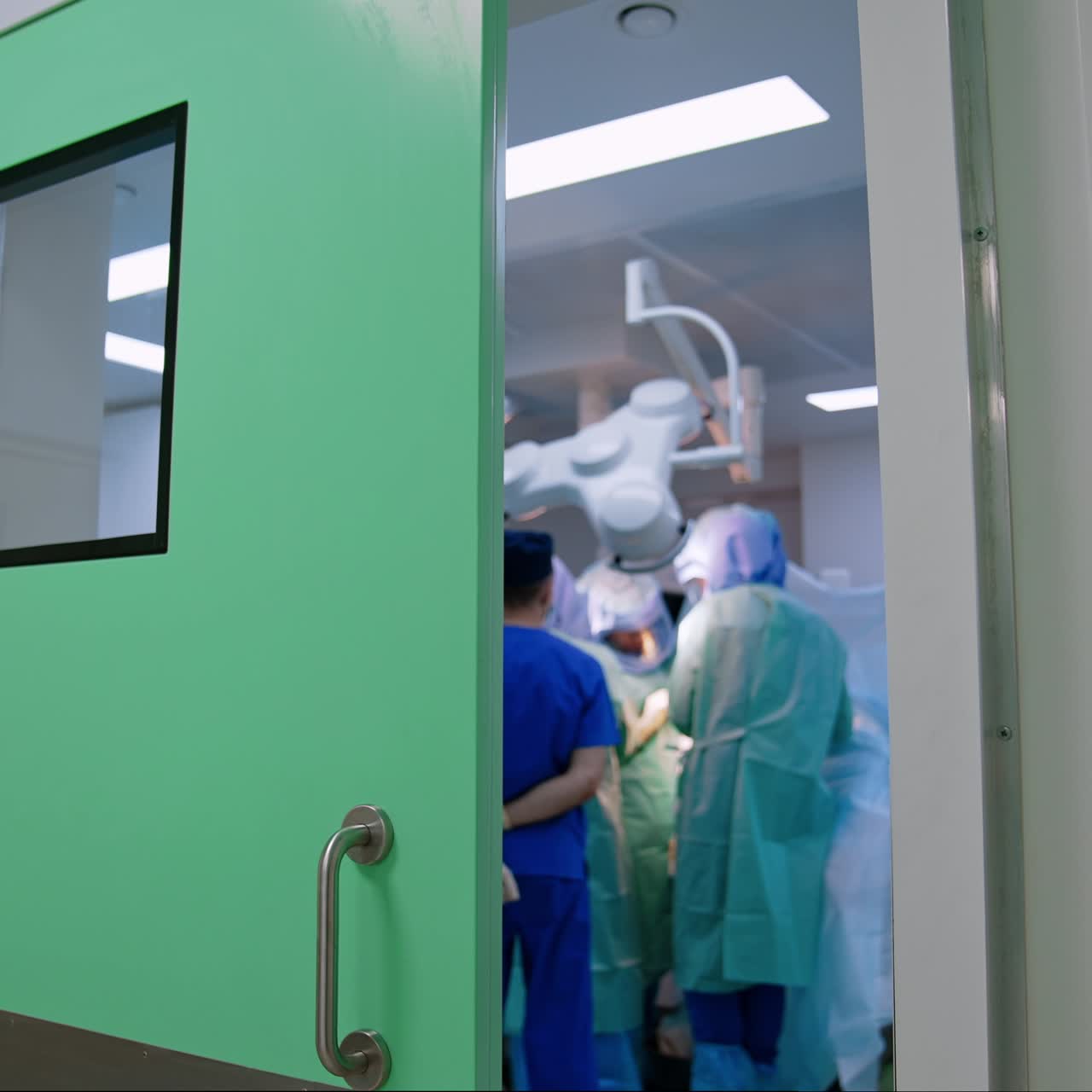 Uma porta grande se abre para uma sala de cirurgia moderna com uma operação em andamento. Médicos estão ao redor da mesa de operação. Um anestesista se movimenta.