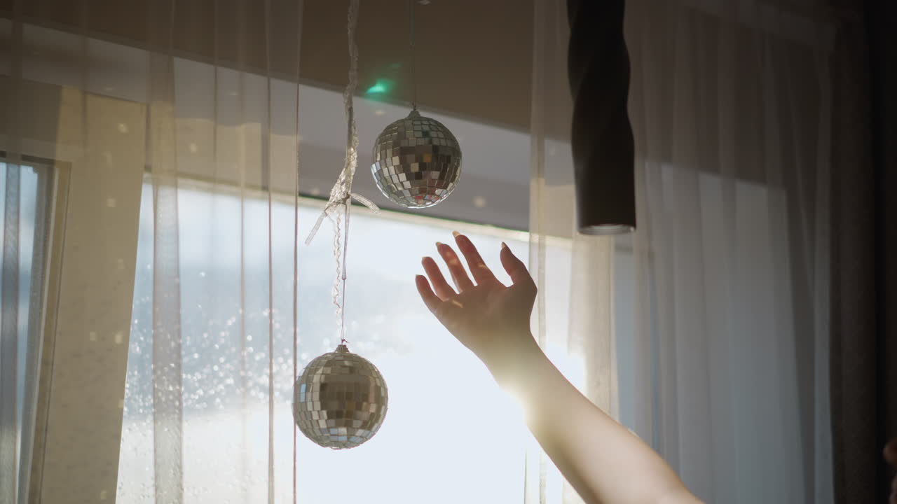Hand touches shiny disco ball decoration hanging near window with sheer curtains while bright sunlight creates glowing reflections and sparkling atmosphere in modern interior home detail scene
