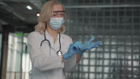 a blonde, middle aged female doctor in a facemask puts on surgical gloves 1