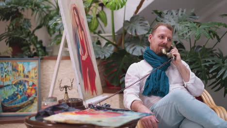 man speaks on phone near portrait on easel. bearded artist sets down palette momentarily to answer important call on retro phone. contemporary art studio