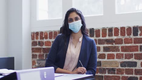 Portrait-of-woman-wearing-face-mask-sitting-on-her-desk-at-office