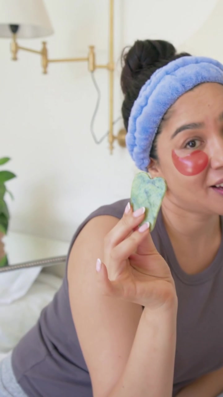 Vertical video: Showing woman holding gua sha receiving device off-screen, teaching skincare on bed