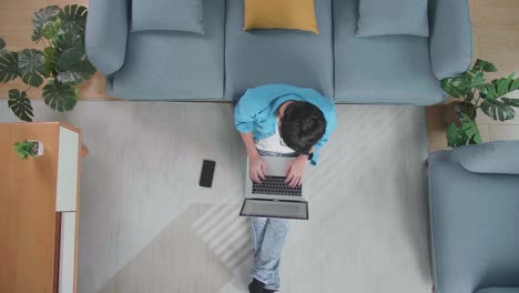top view of a man stretching while typing on a laptop on the floor with smartphone in front of a sofa at home