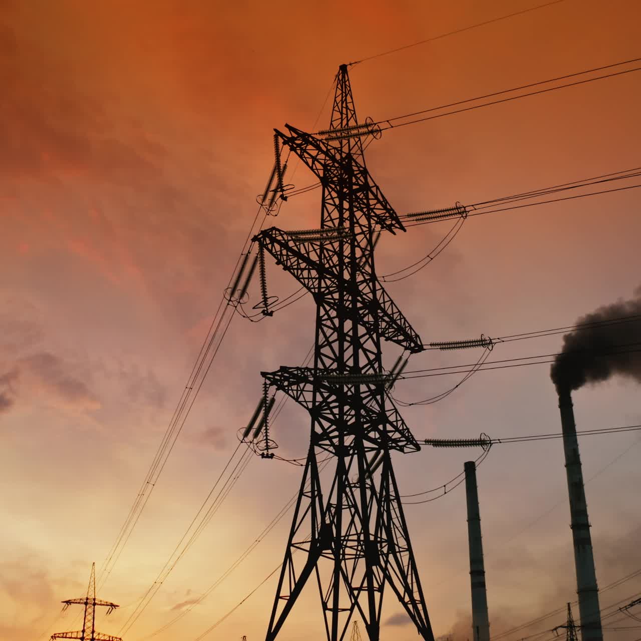 Electric towers near factory chimney with dark smoke. Transmission lines on background of harmful fumes from industry releasing from pipes at sunset. Environment pollution