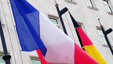 red, white and blue vertical stripes of the french national flag fluttering calmly in the wind on a flagpole next to the german flag in front of a government buidlings in budapest