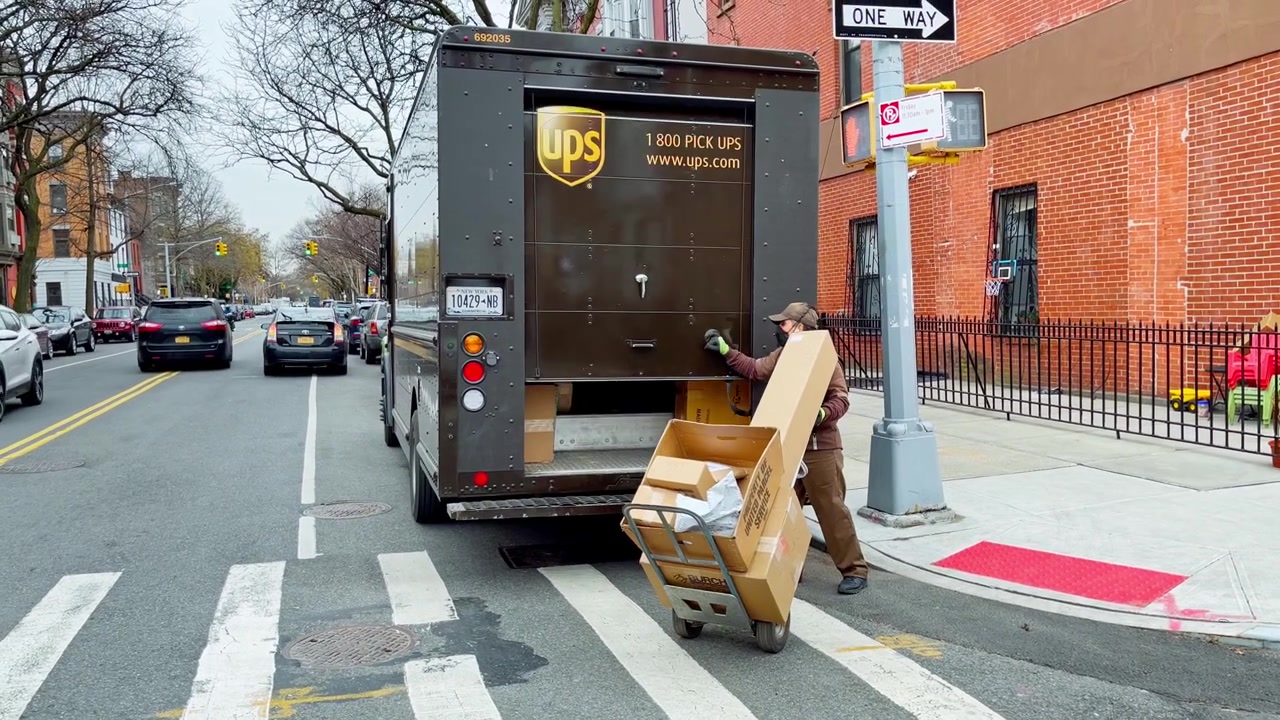 New York, USA, 9 March 2022: Courier unloading cardboard boxes from delivery truck in New York. Delivery worker in face mask moving parcels from brown vehicle to hand truck on New York City street.