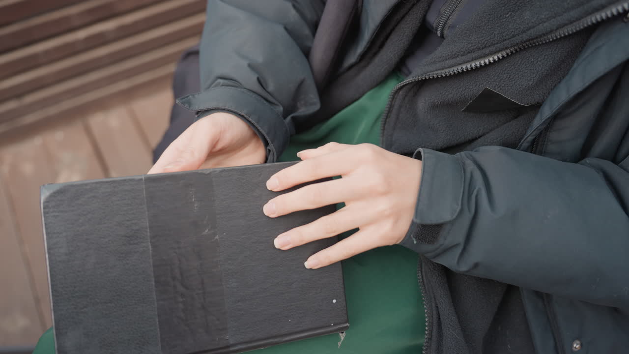 mani che estraggono un libro da una borsa su una panchina. Giovane con giacca che tira fuori un diario usato da una borsa con cerniera, sfoglia le pagine per mostrare testo stampato e foto, primo piano su dita e copertina ruvida, silenzioso