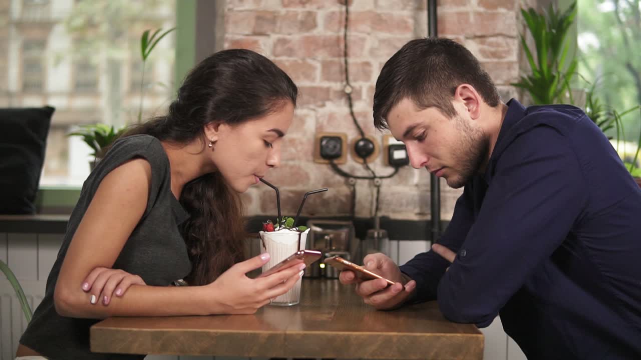 Attractive young couple in cafe using their phones sitting at the wooden table and drinking milkshake using straws. Happy couple