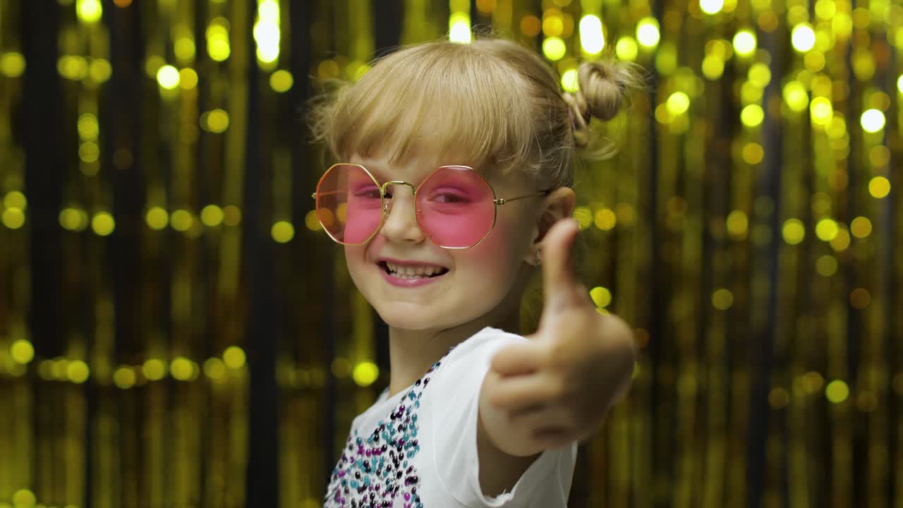 Child show thumbs up, smiling, looking at camera. Girl posing on background with foil golden curtain