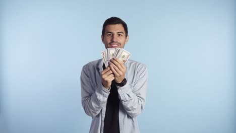 Astonished-caucasian-man-in-casual-clothes-holding-bunches-of-dollars,-lucky-lottery-winner,-super-excited-about-it.-Holding-paper-money-before-face,-can't-believe-in-this-luck.-Blue-wall-background