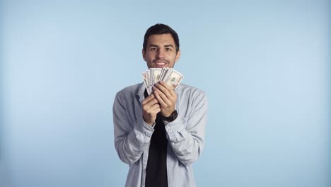 hombre caucásico asombrado con ropa casual sosteniendo montones de dólares, afortunado ganador de la lotería, súper emocionado por ello. sosteniendo dinero de papel delante de la cara, no puede creer en esta suerte. fondo de pared azul