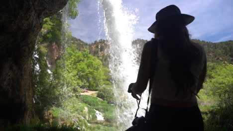 Parte-Posterior-De-Una-Mujer-Caminando-En-Una-Cueva-Bajo-Una-Cascada-Escénica-En-Un-Día-Soleado-De-Verano