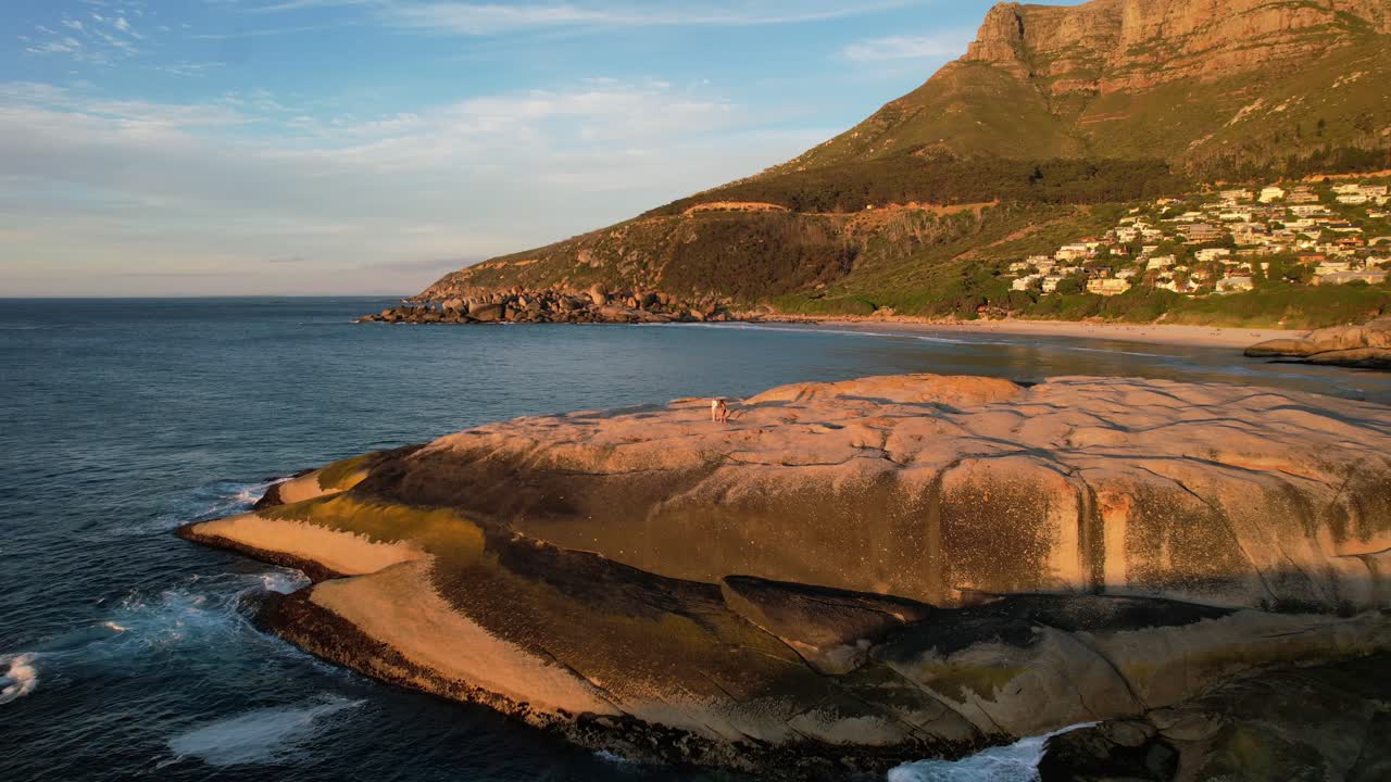 Två flickor går på stora stenblock vid Llandudno Beach med Taffelberget i bakgrunden i Kapstaden under solnedgångens gyllene timme, flygfoto