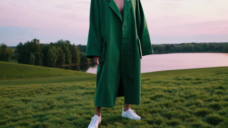 a person in a green outfit and white sneakers walks through a grassy field near a lake at sunset
