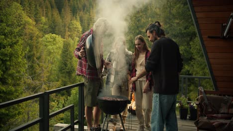 A-group-of-friends-at-a-picnic:-a-guy-in-a-red-shirt-lifts-the-lid-of-the-barbecue-and-thick-white-smoke-comes-out.-Grilling-food-at-a-picnic-with-mountain-and-forest-view