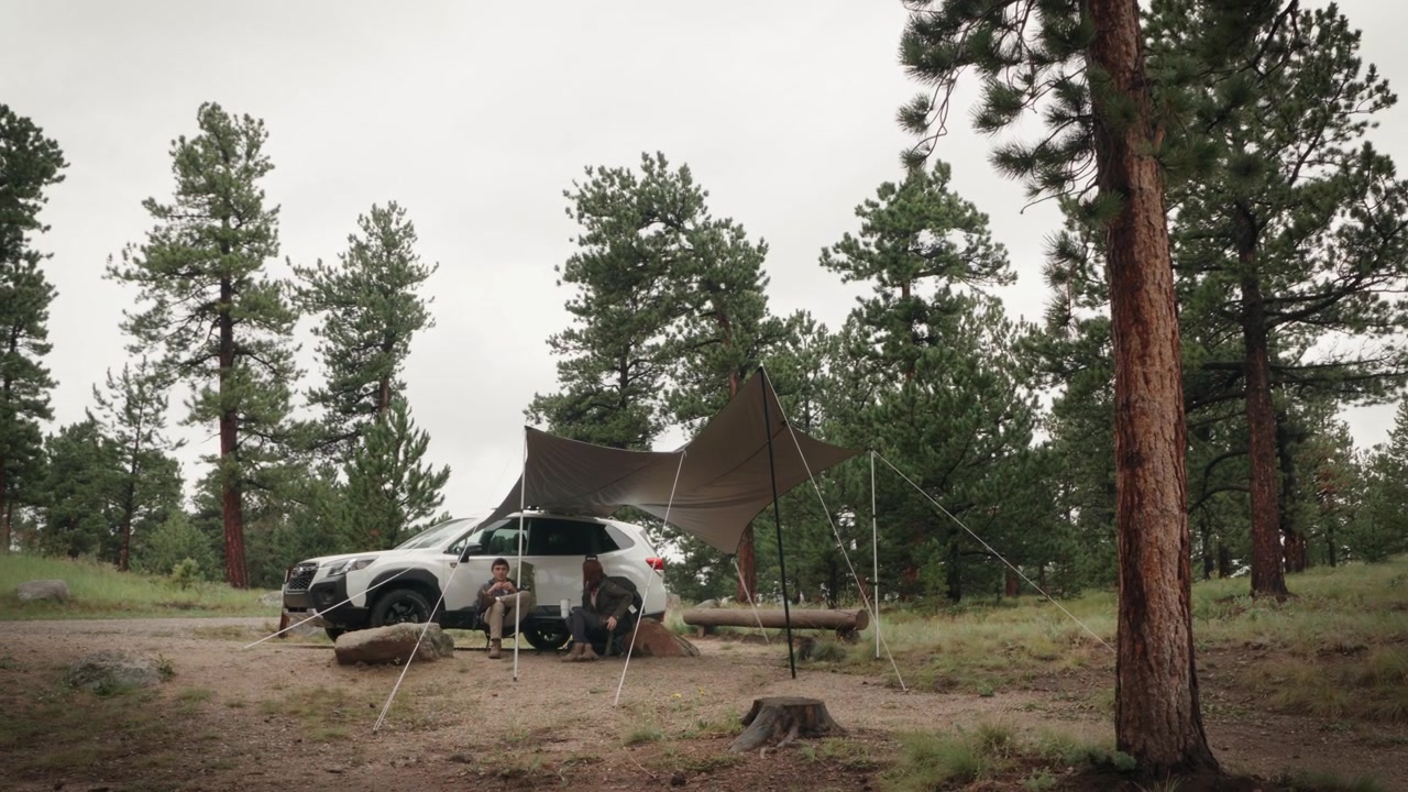 Vidvinkelbillede af en campingplads med bilmarkise i en skov, med et par siddende under den.