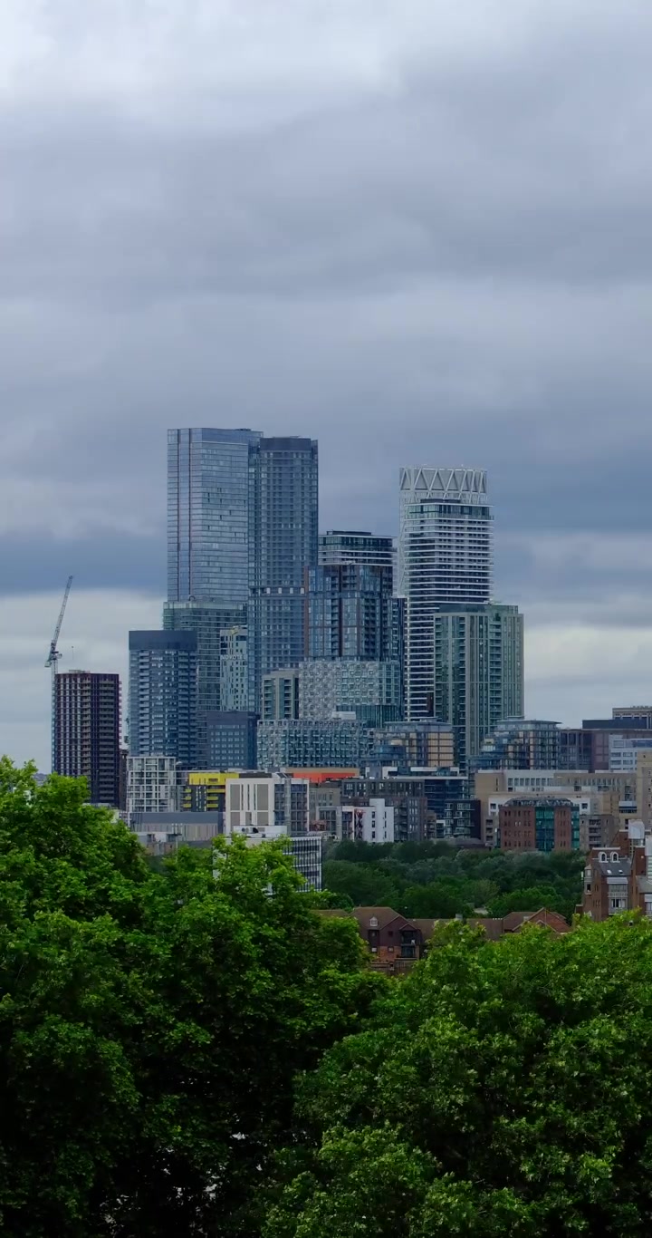 Timelapse vertical dos arranha-céus de Canary Wharf vistos acima dos telhados de Londres e vegetação sob um céu dramático