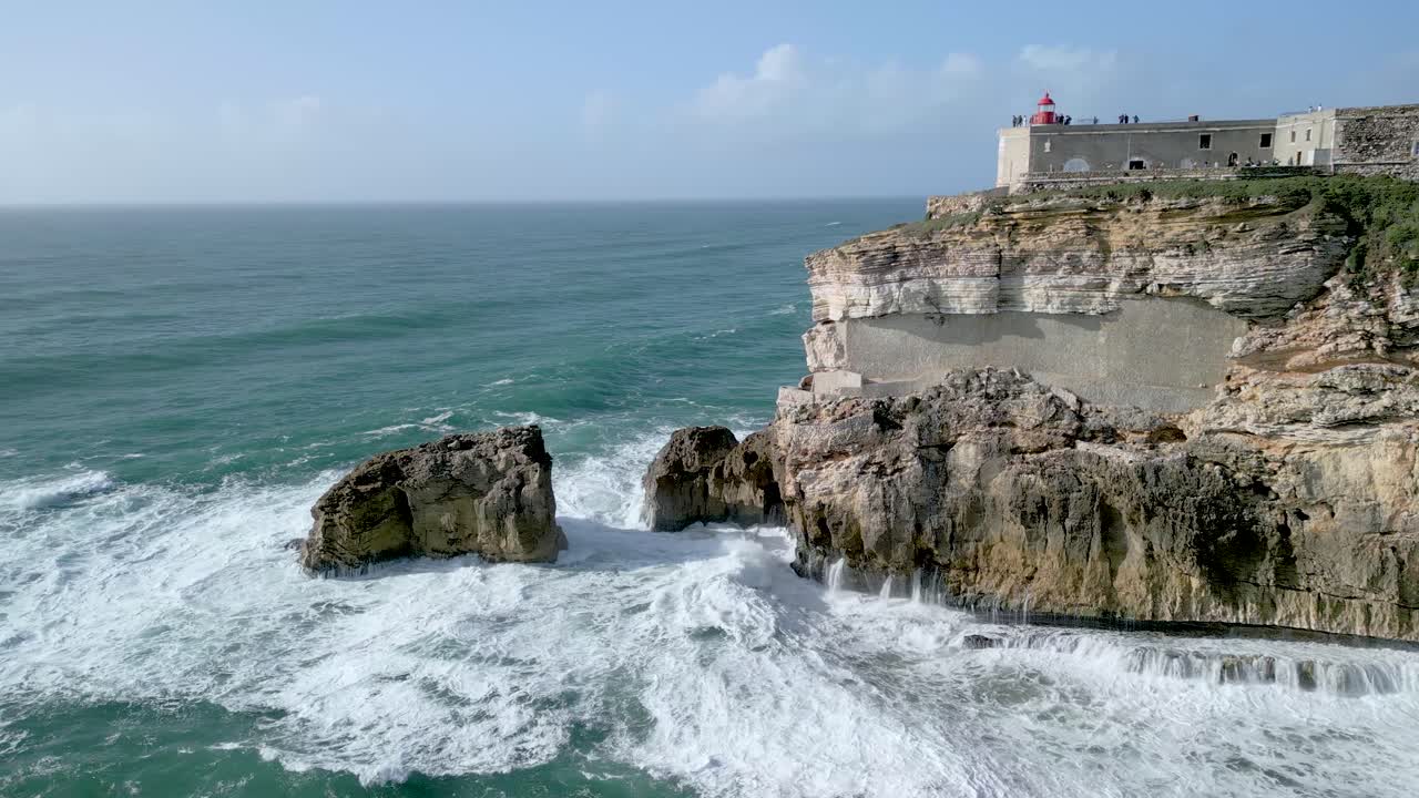 olas chocando contra los acantilados con el fuerte de sao miguel arcanjo en nazare, portugal