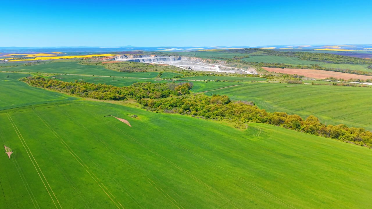Panorama of green agricultural landscape with distant stone quarry. Wide aerial view of vast vibrant green fields under clear blue sky with industrial mining quarry on horizon.