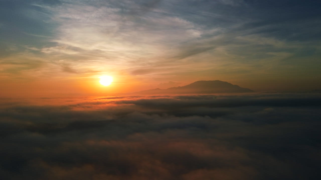 Cinematic drone footage flying over a dense layer of fluffy clouds towards a glowing sun with a mountain silhouette in the distant horizonsunset, clouds, aerial view, sky, sun, horizon, sunrise, heave