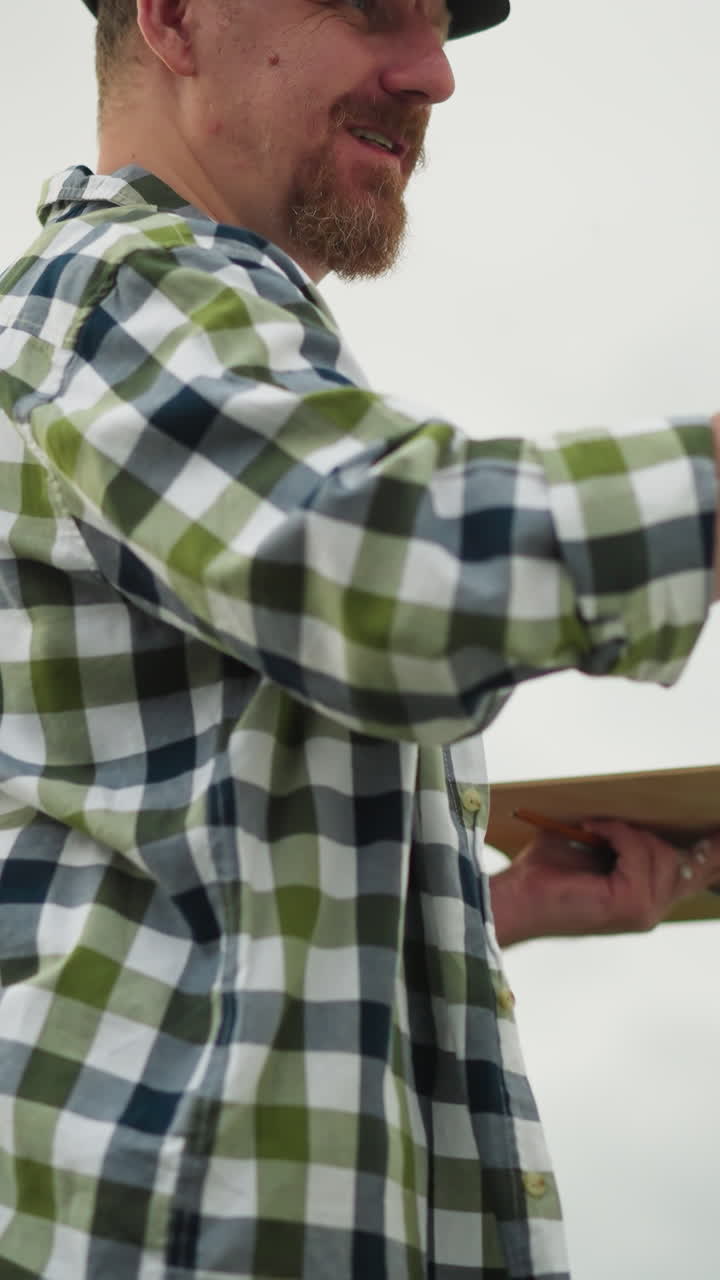 A painter, wearing a black hat and checkered shirt, is captured smiling as he is fully engrossed in his painting. His joy and focus on his creative work are evident in this artistic outdoor scene