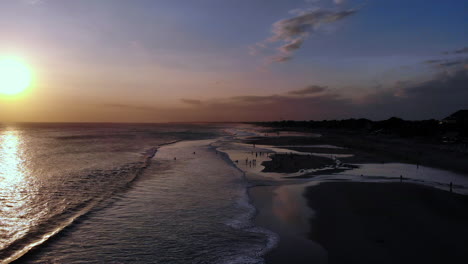 Increíble-Toma-Aérea-De-Una-Colorida-Puesta-De-Sol-En-Una-Playa-De-Arena,-Gente-Jugando-En-La-Playa,-Volando-De-Costado-Hacia-El-Océano-Desde-La-Playa