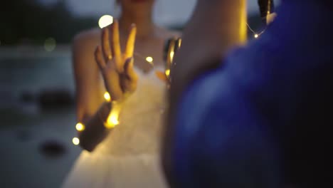 novia y novio parados juntos en la playa con copas de vino y guirnaldas de luces