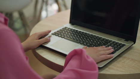 young woman in pink shirt focuses on her laptop on a wooden table, as she opens it while sitting in a restaurant, blurred chair is seen by the side and lights create a soft bokeh background