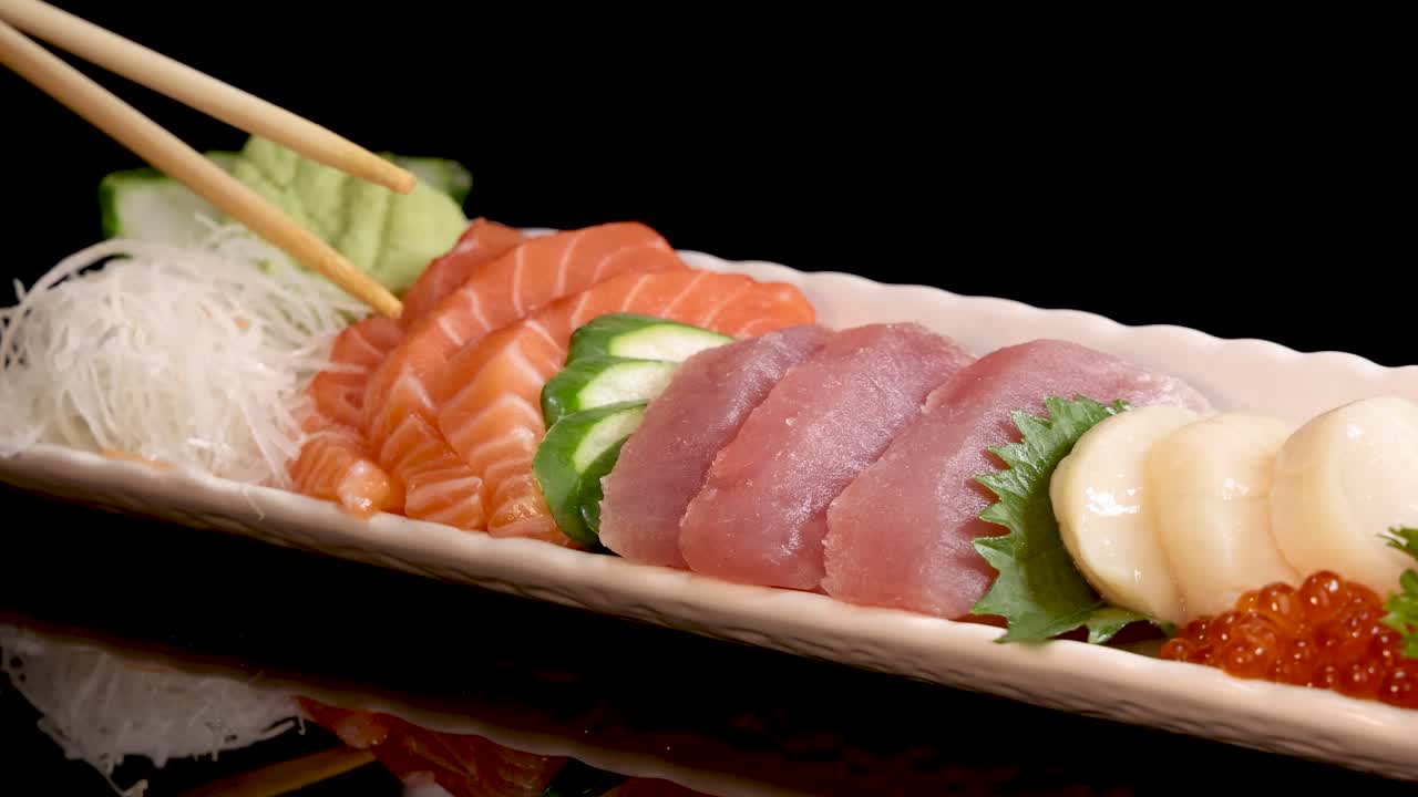 Chopsticks lift a slice of tuna sashimi from an assorted Japanese sushi platter with salmon, scallop, roe, and garnishes on a black background under studio lighting