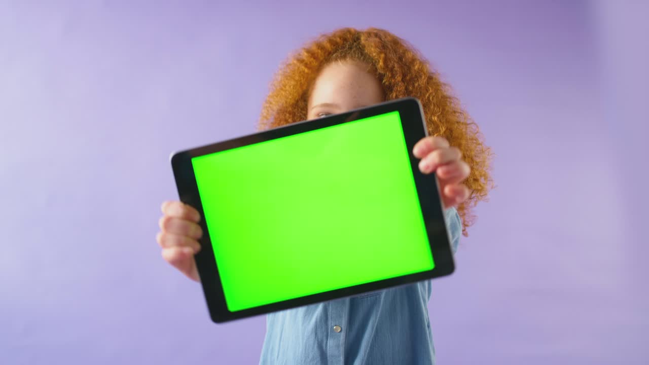 Studio shot of smiling girl holding digital tablet with green screen looking at camera against purple studio background - shot in slow motion