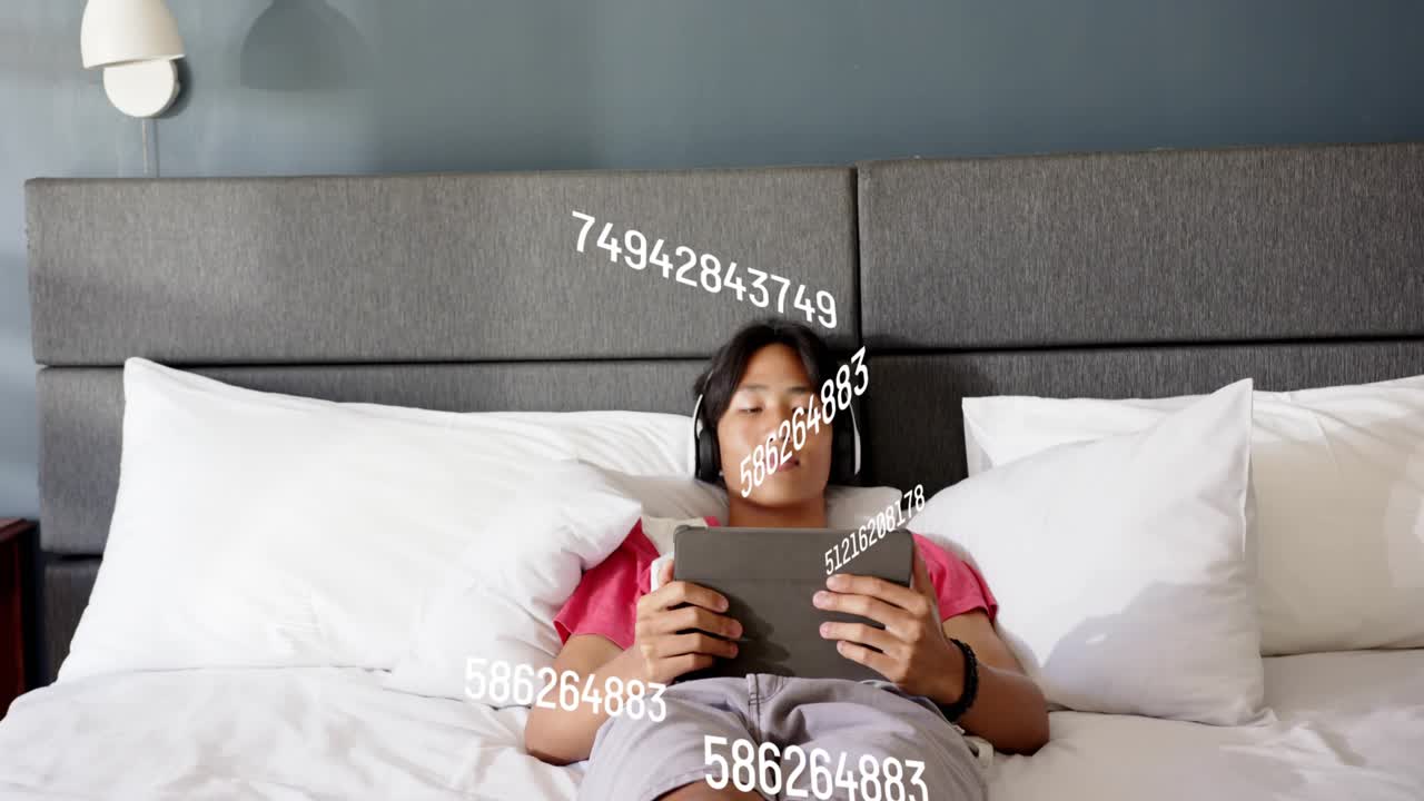 Male teen tapping tablet in bed, causing number overlay orbiting it, reclining, viewing tech data