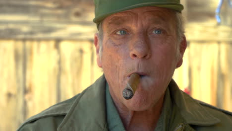 closeup of male with gray hair wearing army clothes, puffing smoke from his cigar, looking around confidently