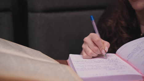 close up of female hand holding pen writing inside pink notebook filled with text while old brown book lies open on wooden table, creating academic mood in cozy indoor space with soft lighting