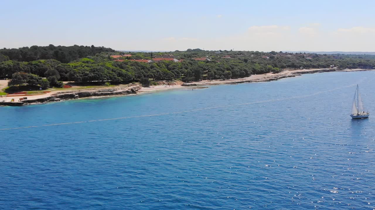 Flight over white sailboat sailing along a coast under blue skies on calm clear blue water. Tracking shot