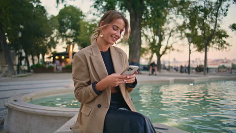 carefree lady enjoy chatting smartphone sitting at water fountain in evening.