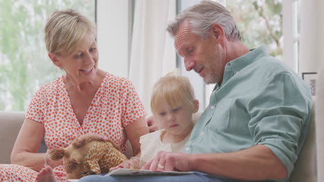 Abuelos-Sentados-En-El-Sofá-Con-Su-Nieta-En-Casa-Leyendo-Un-Libro-Juntos