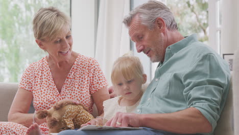grandparents sitting on sofa with granddaughter at home reading book together