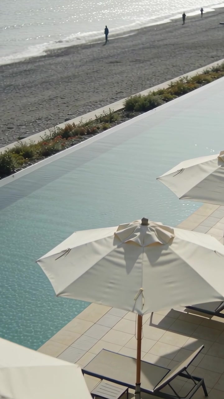 Vertical video: Framing sun umbrella at resort pool, two pedestrians walking shoreline admiring sea