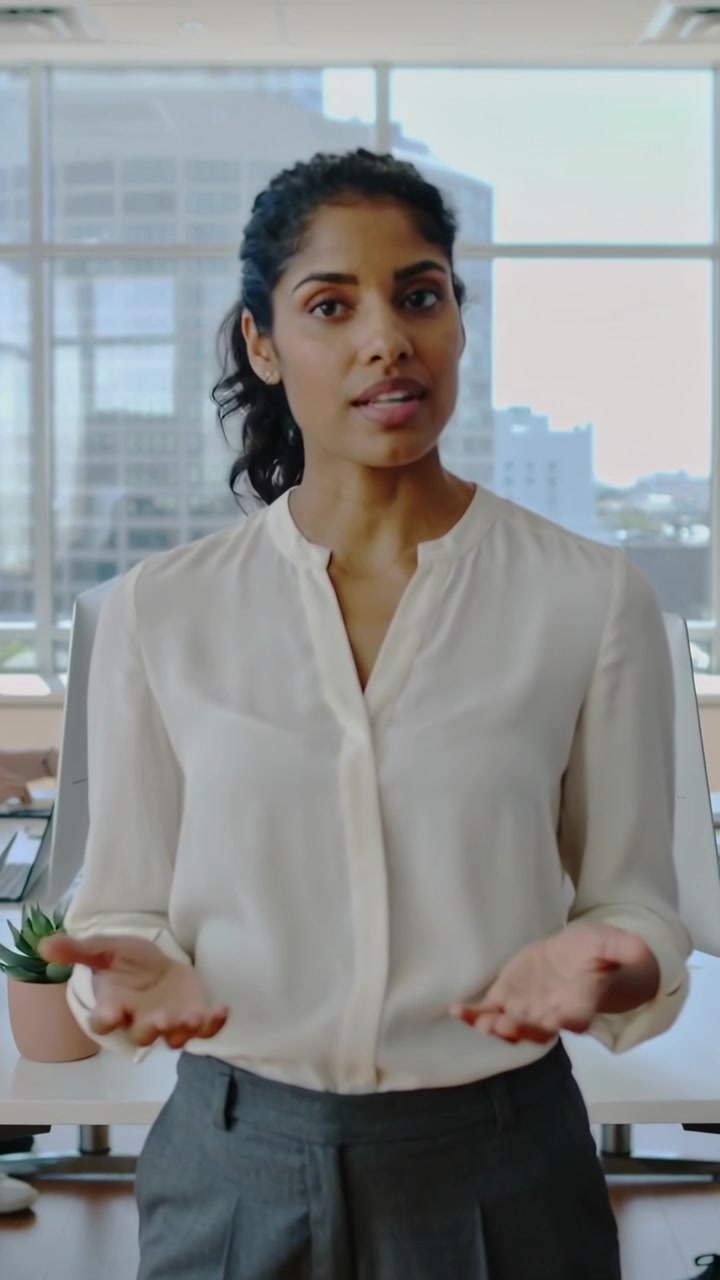 Vertical video: Camera moving closer, woman in business casual presenting, gesturing at office desk