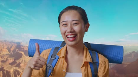 close up of asian female hiker with mountaineering backpack smiling and showing thumbs up gesture to camera while traveling at the top of mountain