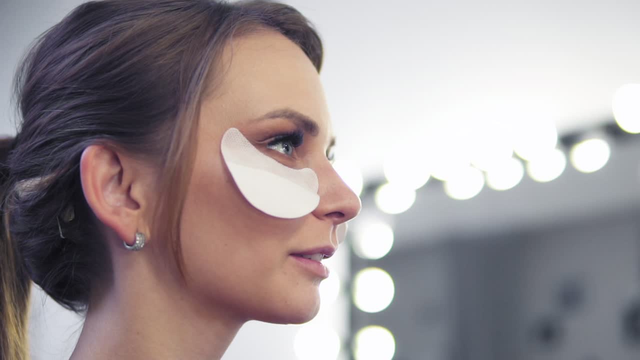 젊은 모델이 메이크업 아티스트가 눈꺼풀에 눈조림을 바르고  ⁇ 색 패치로 피부를 보호하기를 기다리고 있습니다. 전문 메이크업