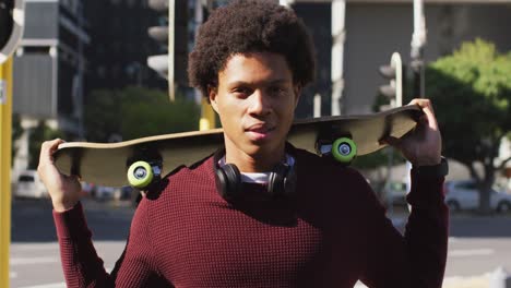 Portrait-of-african-american-man-in-city,-standing-holding-skateboard-on-shoulders-and-smiling