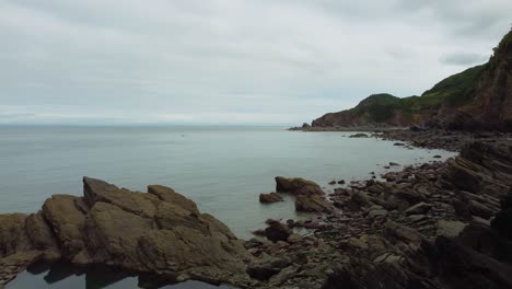 Aerial-Drone-Panning-Shot-of-Dramatic-Rocky-Cliffs-Trees-and-Sea-with-Kayaks-Woody-Bay-North-Devon-UK-4K