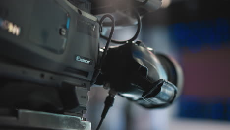 close up camera lens mounted on professional video rig with attached viewfinder and cables reflecting blue studio background lights, highlighting metallic textures and precision focus