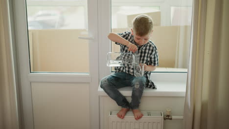 child seated on windowsill pouring water from large transparent jar into glass cup placed on leg, wearing checkered shirt and jeans, carefully concentrating indoors on bright sunny day